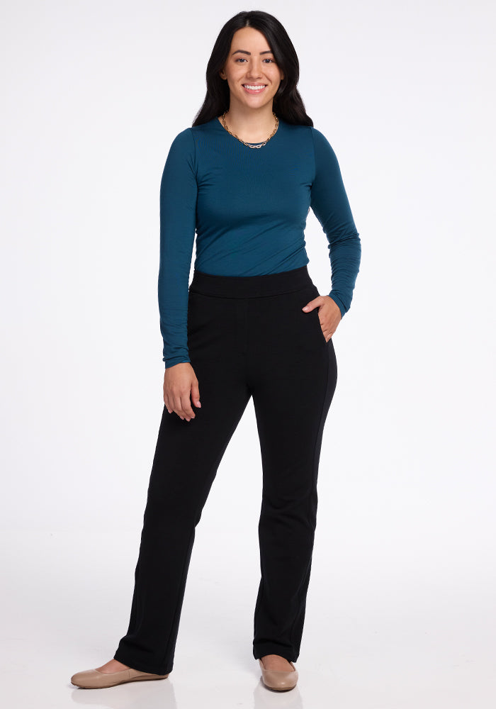 A person with long dark hair is smiling and standing against a plain white background. They are wearing a teal long-sleeved top, Emerson Straight Leg Pants Petite - Black from Woolx, which are made of moisture-wicking fabric, and beige shoes. One hand is relaxed by their side, and the other is in a pocket.