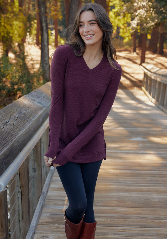 A woman with long brown hair smiles on a sunlit forest bridge, wearing the Woolx Evie Ribbed Tunic, dark leggings, and brown boots.
